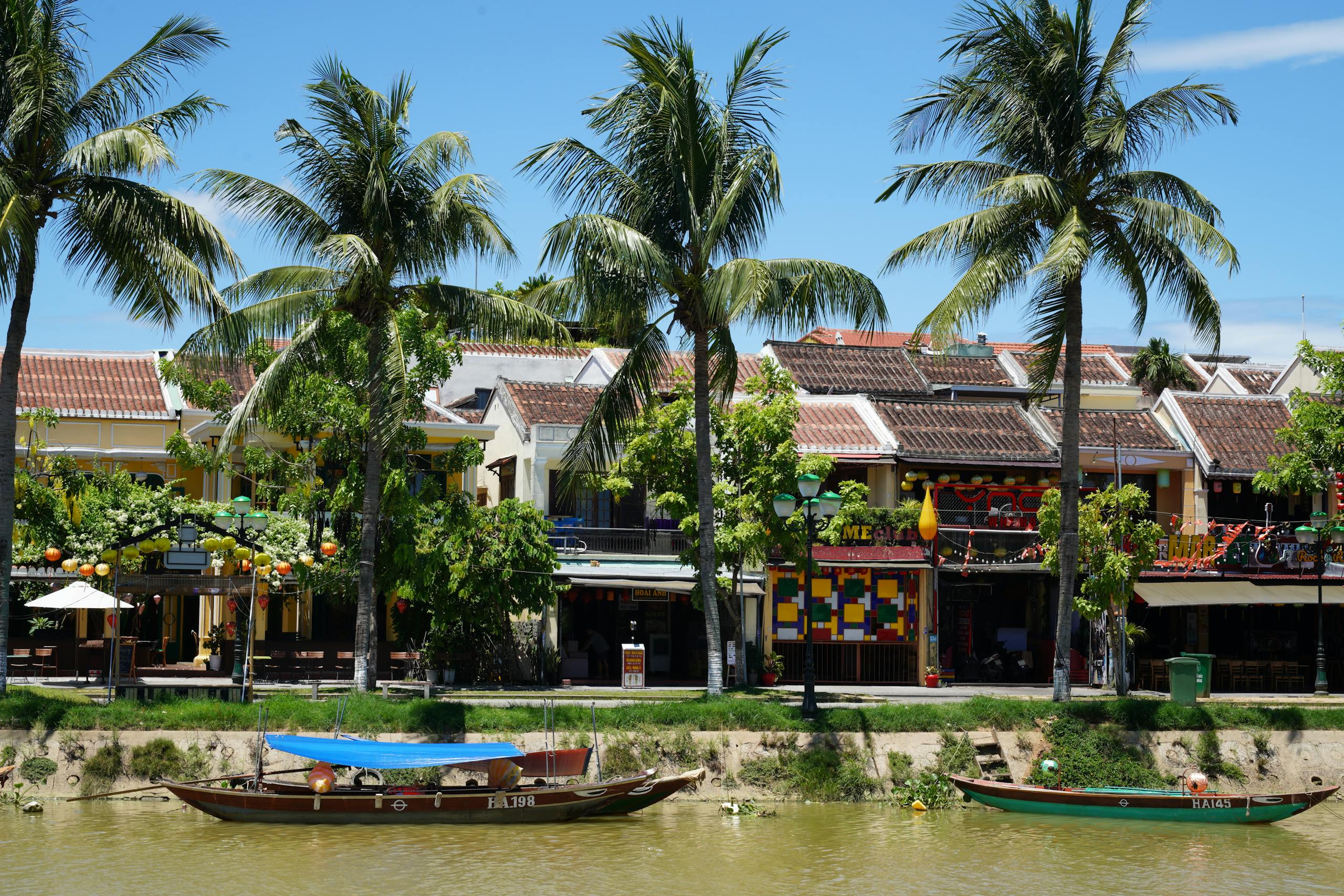 Colorful riverside town with lush palm trees and quaint boats, under a bright blue sky.