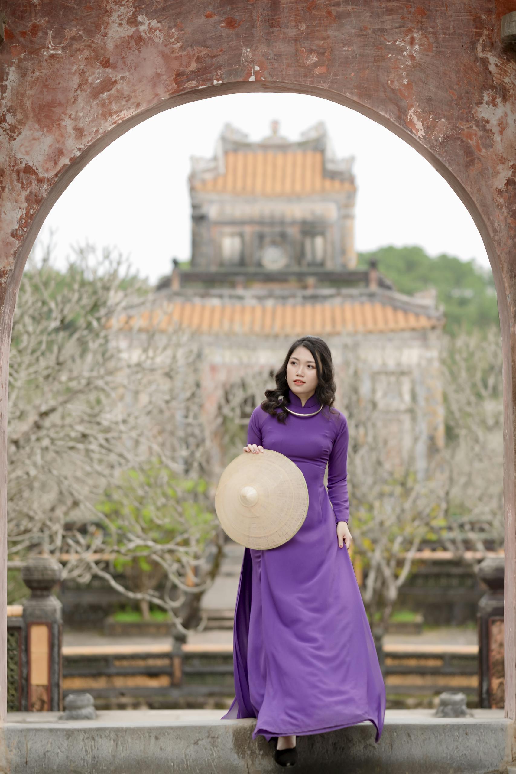 Asian woman in purple dress holding conical hat, standing at historical site.