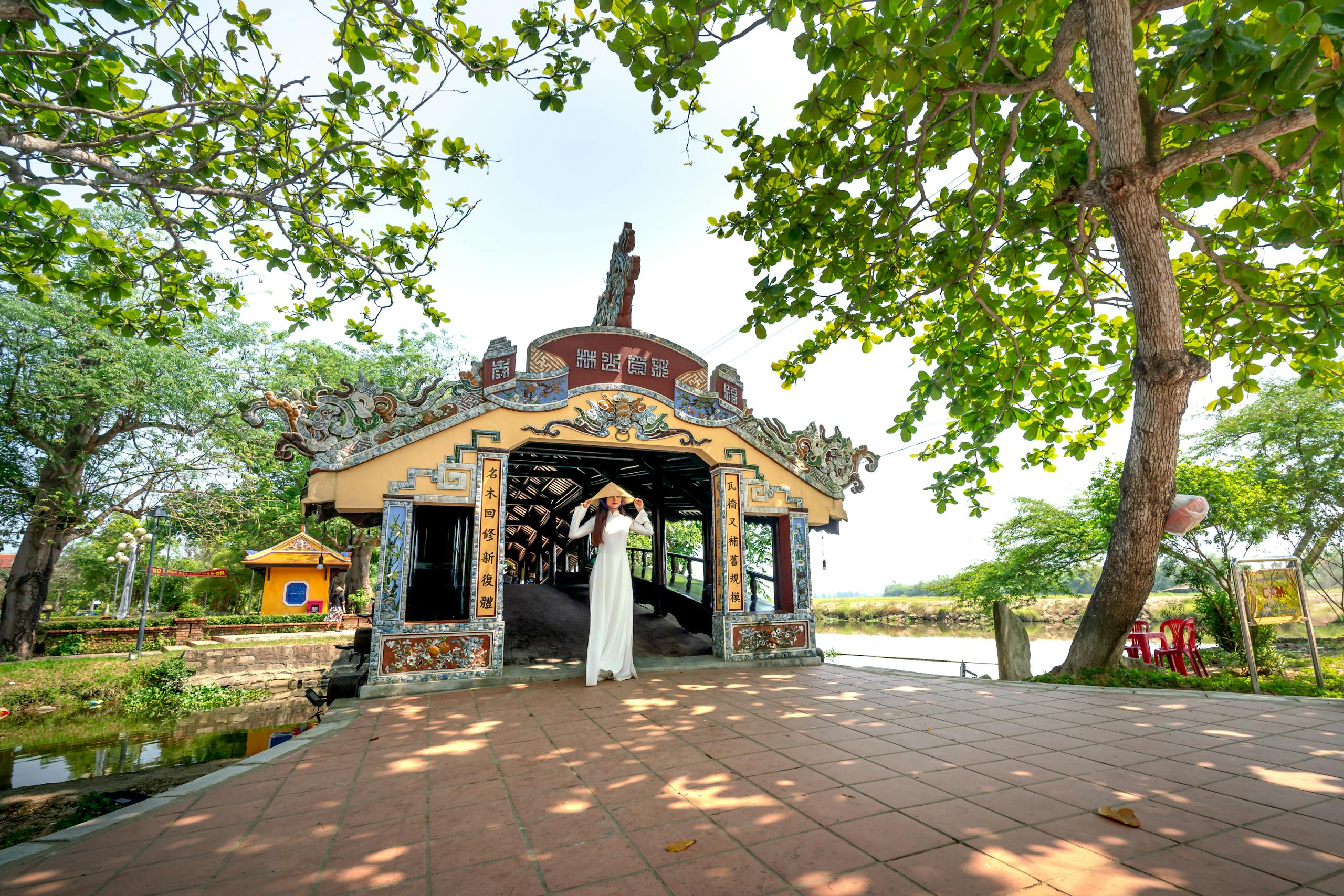 Top Day Trips from Hue: Discover the DMZ, Lang Co Beach & Bach Ma National Park 9 Elegant Asian woman wearing ao dai, posing on historic Thanh Toan Bridge in vibrant outdoor setting.