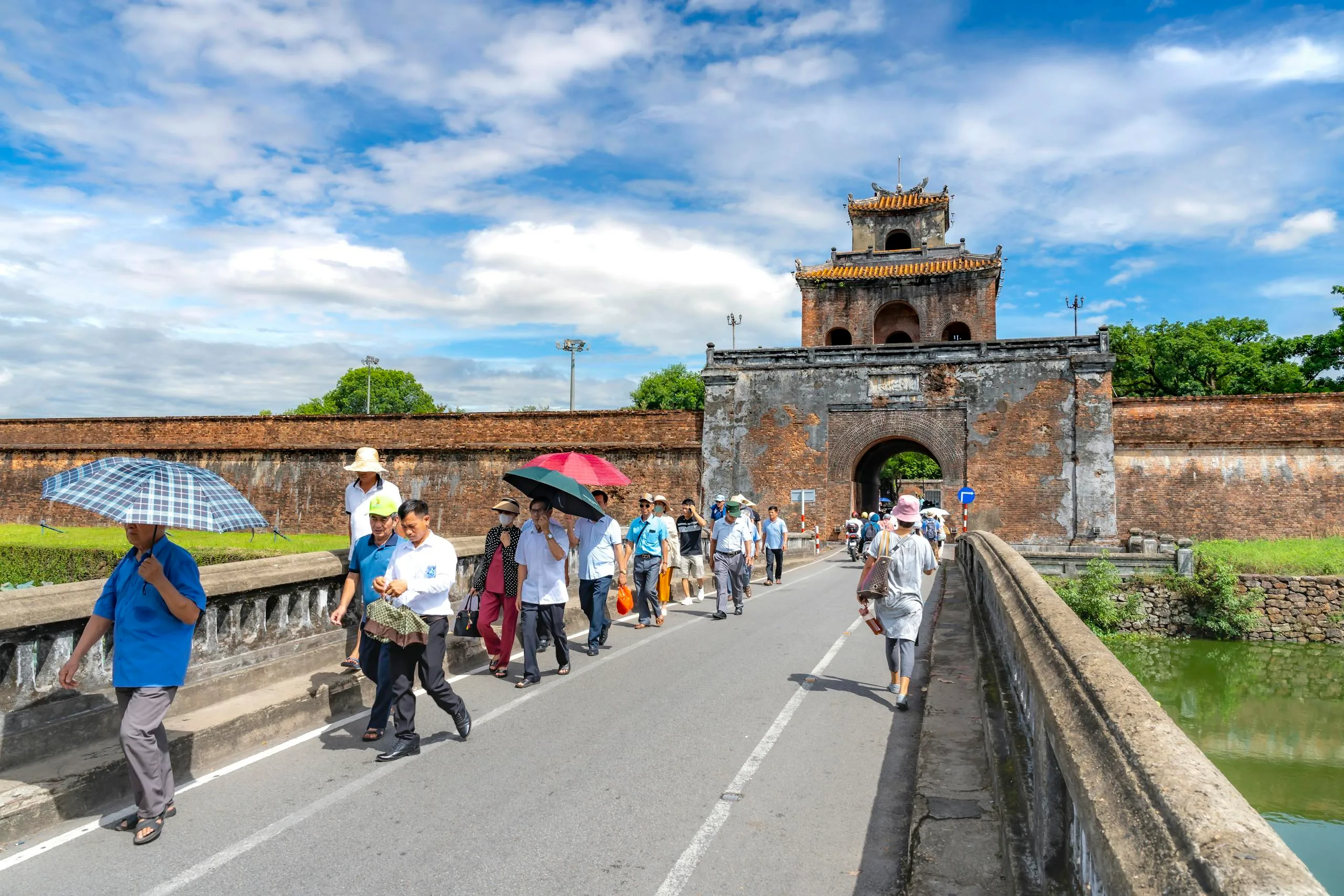Tourists stroll by ancient gate at Hue Citadel under bright blue sky in Vietnam.