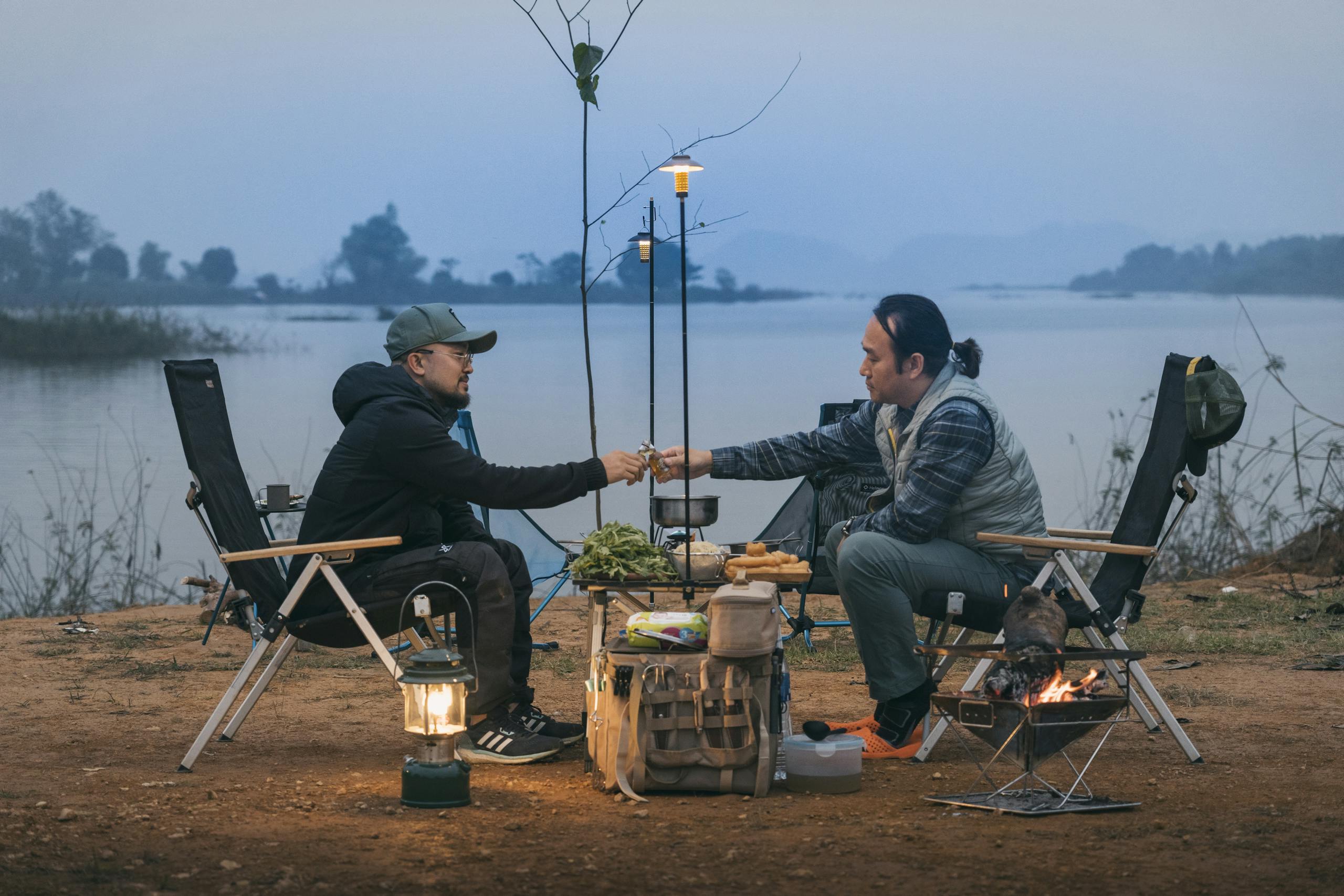 Two men enjoying a campfire by a lakeside in Sơn Tây, Vietnam during dusk.